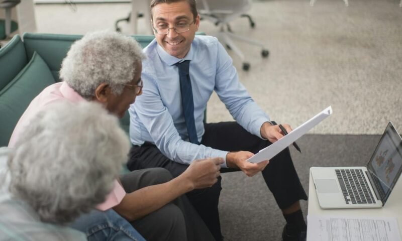 Consultant discussing financial plans with senior clients in a modern office setting, using documents and a laptop.