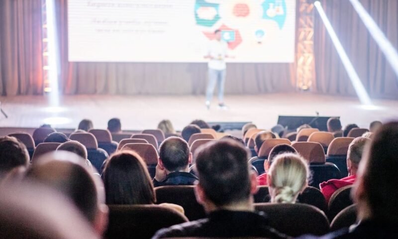 Audience attentively watching a presentation in a conference setting, with stage lights and speaker visible.