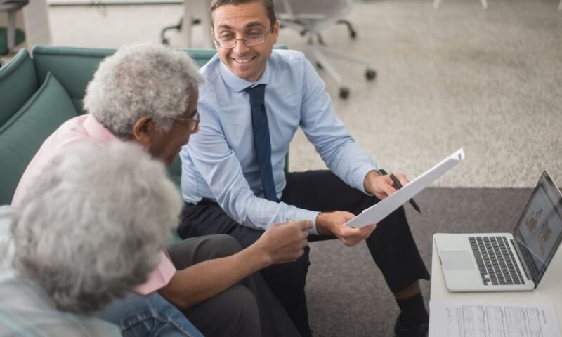 Consultant discussing financial plans with senior clients in a modern office setting, using documents and a laptop.