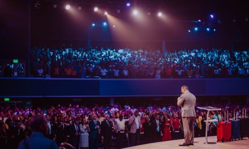 A speaker in a gray suit captivates a large crowd on stage during a motivational event.
